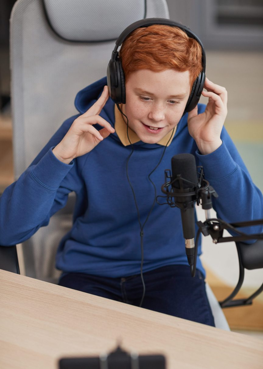 Vertical high angle portrait of red haired teenage boy speaking to microphone and recording video while streaming online
